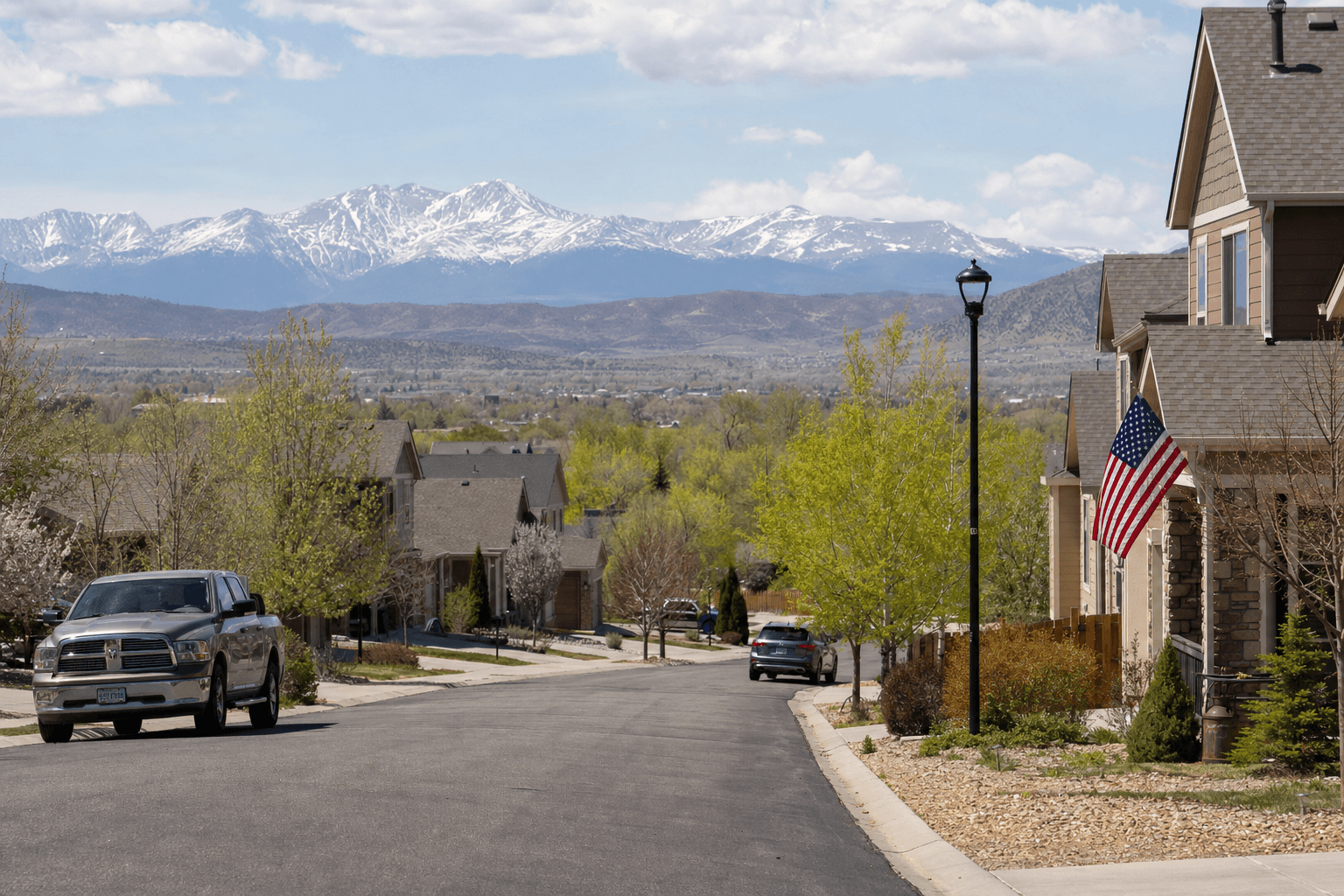 Residential street with houses and parked cars, budding spring trees, an American flag on a porch, and snow-capped mountains on the horizon.