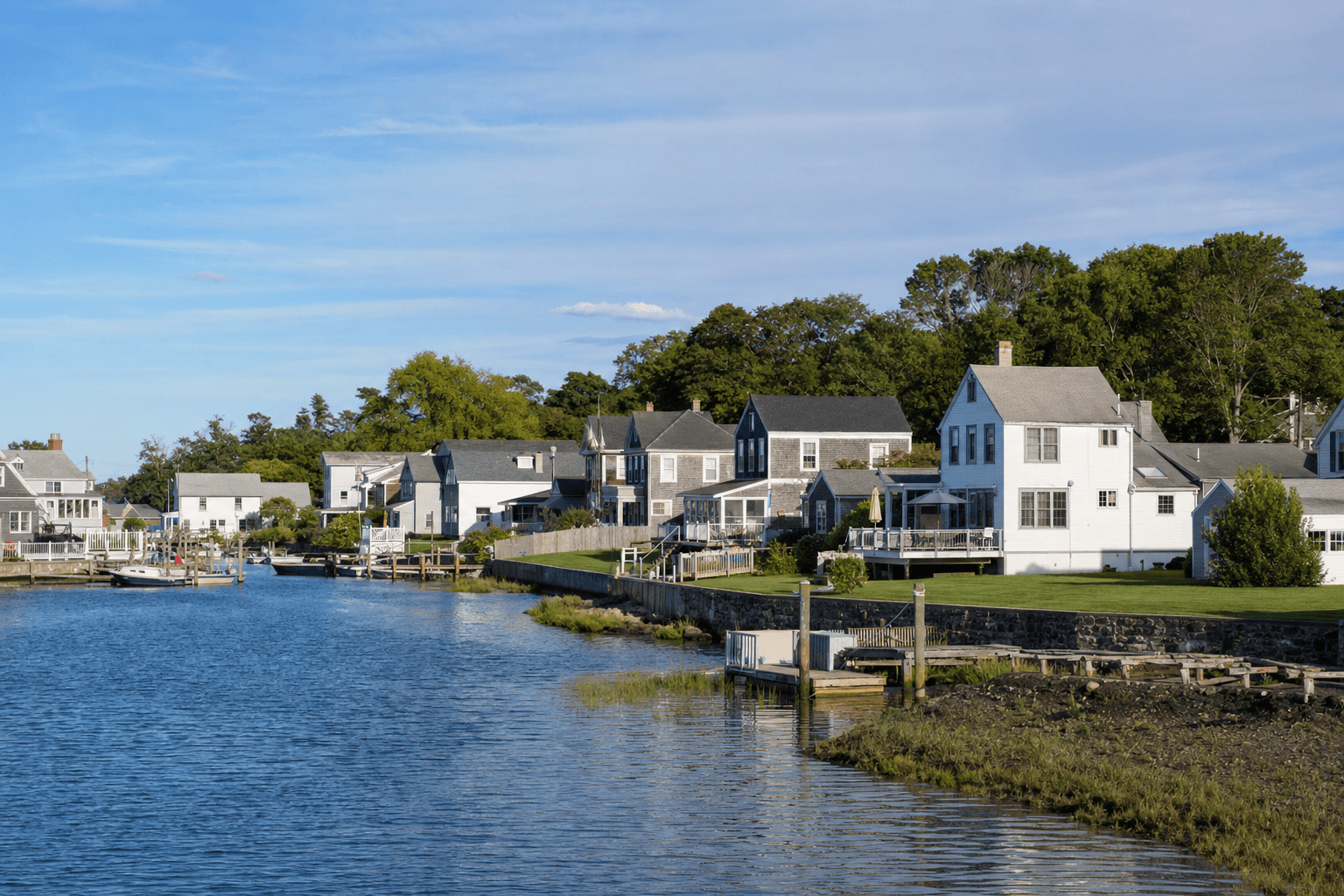 Clapboard and shingle waterfront houses and small docks lining a calm tidal inlet with boats and trees under a clear blue sky