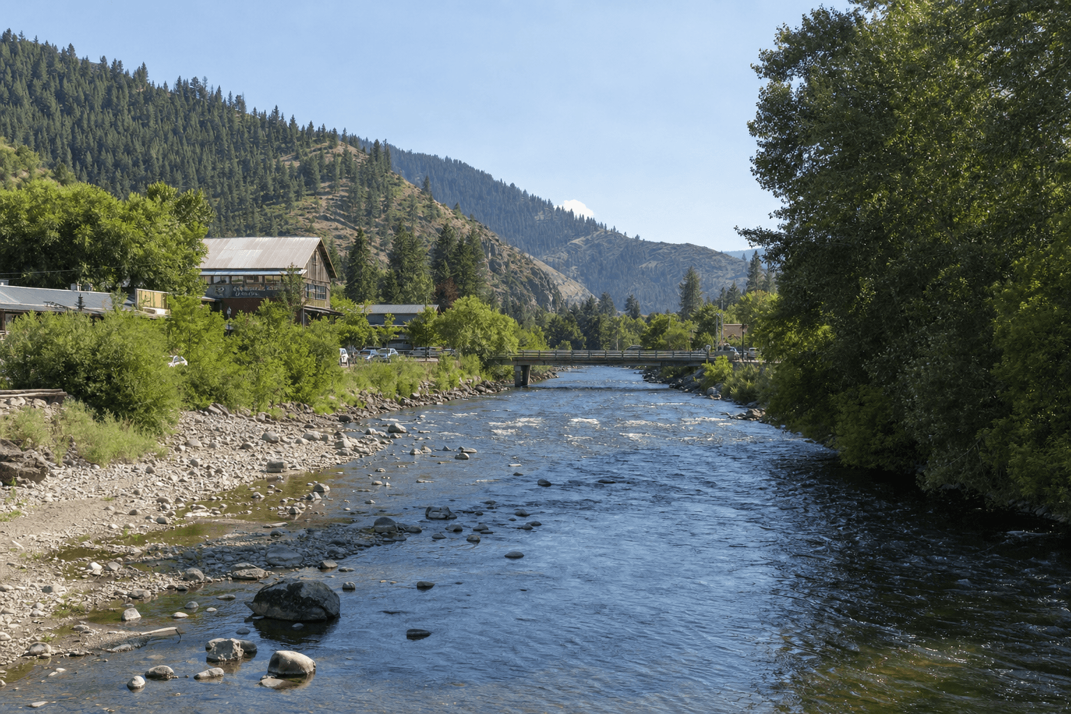 River flowing over rocks toward a low bridge, with leafy trees and a rustic riverside building along the bank and forested mountains rising in the background under a clear sky.