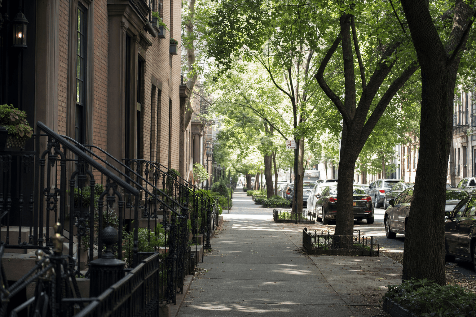 Shaded, tree-lined sidewalk flanked by brownstone stoops, wrought-iron railings, and parked cars.