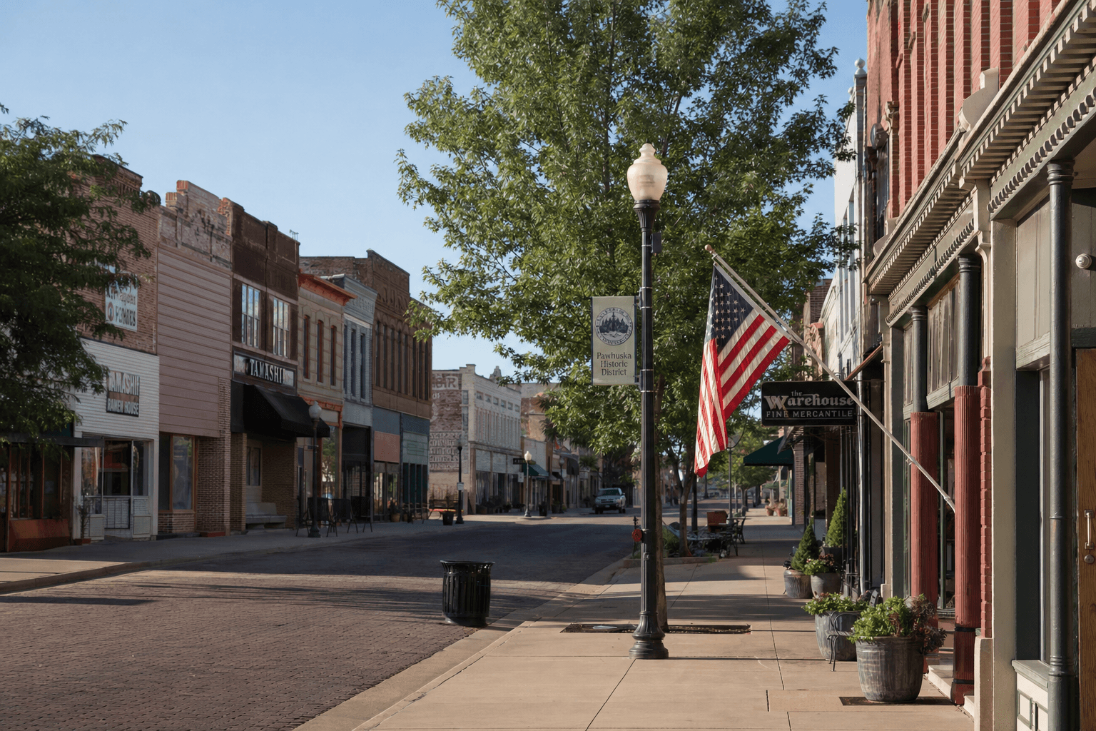 A quiet small-town main street lined with brick storefronts, a lamppost with an American flag, and trees shading the sidewalk.