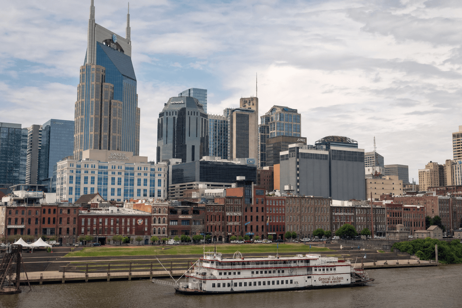 A large white paddlewheel riverboat named General Jackson docked on the river in front of a row of brick waterfront buildings with modern skyscrapers rising behind them under a cloudy sky.
