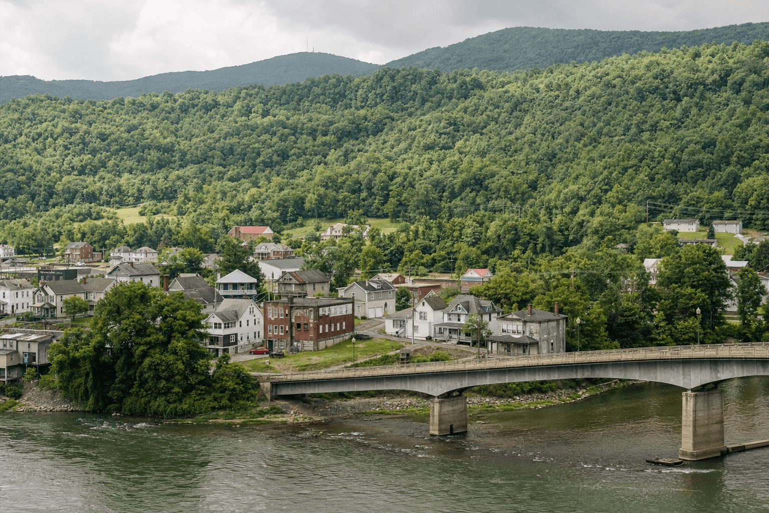 A small riverside town with a concrete bridge crossing a calm river and tree-covered hills rising behind rows of houses.