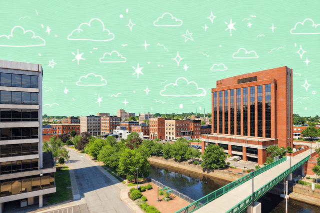 Panoramic view of downtown Bangor, ME, featuring a river, red brick buildings, modern offices, and a pedestrian bridge under a bright blue sky.